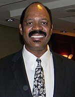 A black person, wearing a black suit and a tie, is posing for a photo.