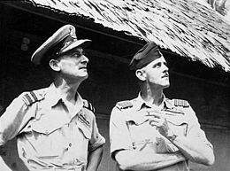 Half length portrait of two men in short-sleeved tropical military uniforms with pilot's wings, standing in front of a hut and looking into the sky. One is shaven and wears a peaked cap, with hands on hips. The other has a moustache and wears a forage cap, with arms crossed and a cigarette in his left hand