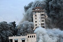 Two buildings are enveloped in clouds of black smoke with one damaged and falling.