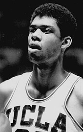 A young black man is completing a two-handed reverse slam dunk during a college basketball game. The photograph is in black and white.