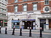 Across a street and behind black bollards is a brick building of at least two storeys. The ground floor is stone coloured and two people are standing in a dark entrance beneath a blue rectangular sign reading "MOORGATE STATION" in white. Above this, attached to be wall at 90 degrees is a white rectangular sign with the National Rail logo and London Underground roundel. Above this, below three windows, blue lettering reads "MOORGATE STATION"