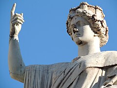 Haut d'une statue blanche, on voit la tête et un bras, un doigt pointant vers le ciel.