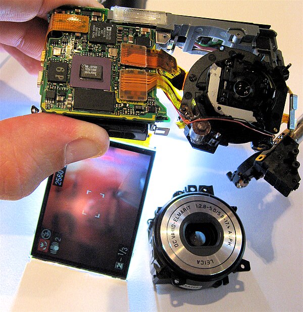 Digital camera, partially disassembled. The lens assembly (bottom right) is partially removed, but the sensor (top right) still captures an image, as 