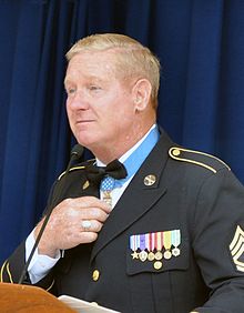Head and shoulders of a blond-haired white man in military uniform, standing before a microphone. His right hand is touching a medal hanging from a light blue ribbon around his neck.