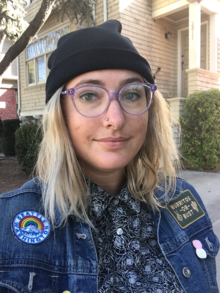 Portrait of a white woman with blonde hair. She is wearing a black beanie and a jean jacket.