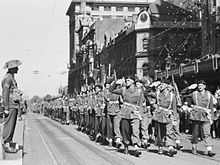 Black and white photo of soldiers wearing military uniforms marching in a close formation down an urban street. The soldiers at the front of the photo are saluting. Another soldier is standing at attention at the left of the photo.