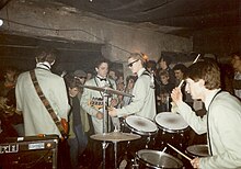 A rock band, the U-Men, playing onstage in a small venue with low ceilings. The band members are wearing matching grey suits and bow-ties.