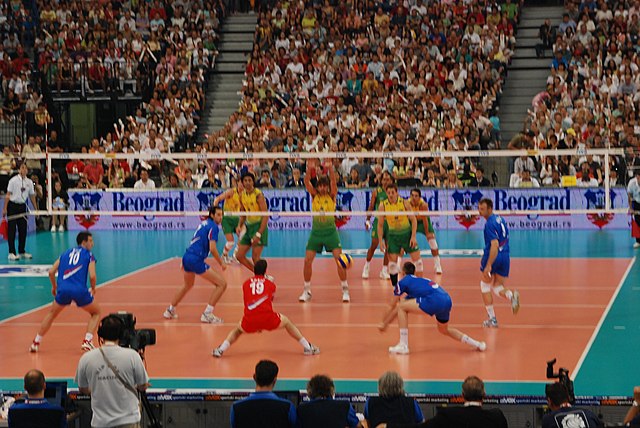 vue de derrière d'un mathc de volley-ball: au premier plan, la Serbie, avec un libéro en rouge qui s'apprête à réaliser une réception