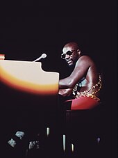 An adult male with sunglasses plays a piano under a spotlight on a darkened stage, 1973