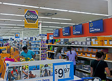 Several people shopping in an area with high shelves on the right stacked with spiral notebooks and other stationery products in open yellow boxes. At the top of the shelves are several blue signs with a small stylized starburst logo in yellow and "Everyday Low Price" in white text, on a red background. Strip fluorescent lights on the ceiling illuminate the scene; a yellow sign hanging from the ceiling has an octagon with "back to school" and text in English and Spanish beneath it. On the left are shelves reaching camera height; a sign in the front bottom says "$9.97".