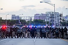 Plusieurs policiers armés en uniforme avec casques et boucliers sur une autoroute barrée, plusieurs édifices et des espaces verts en arrière-plan.