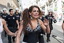 Une femme trans avec une expression de détresse marche dans la rue, suivie d'une dizaine de policiers