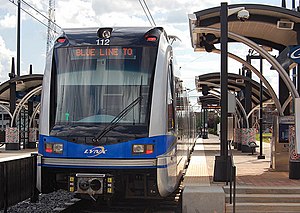 A blue and gray train stopped at a side platform station with station black and gray station canopies visible.