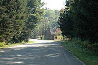 A road through a forest leads to a fieldstone building with a chmney and steep roof next to a stop sign
