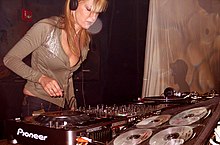 Langhaarige, blonde Frau in tief geschnittenem Pullover und tief geschnittenen Jeans mit Kopfhörern steht vor einer Studio-Soundkarte, die die Schalter einstellt.