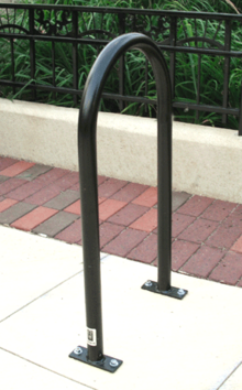 Um tubo de metal pintado de preto dobrado em forma de U e aparafusado em ambas as extremidades a uma laje de concreto, na frente de um caminho de tijolos e grades de ferro