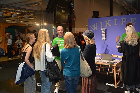 En volontär i grön t-shirt pratar med fyra besökare i Wikipediamontern på Bokmässan.