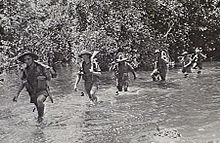 Soldiers wearing slouch hats and shorts wade along a watercourse