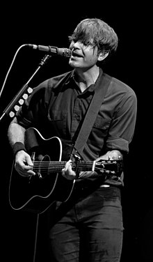 A man sings into a microphone while playing an acoustic guitar.