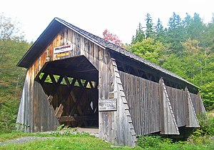 Eine braune Holzbrücke mit Spitzdach. Über dem Portal befinden sich Schilder mit der Aufschrift 