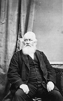 John Cracroft Wilson sentado em uma cadeira, visível de cima da perna, usando medalhas presas ao paletó, com uma barba branca.