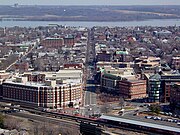 Photo of many buildings. Many are several stories high with red brick facade. They are arranged along a major street which extends from the front of the photo straight toward the back. In front is a Metrorail station with train tracks extending from the left to the right.