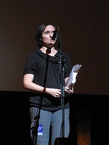 Vowell standing onstage in front of a microphone holding papers