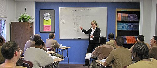A woman standing at the front of a classroom pointing to fractions on a whiteboard. There are eleven men picture sitting at desks in front of her
