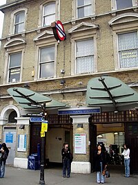 A beige-bricked building with a rectangular, dark blue sign reading "WHITECHAPEL STATION" in white letters all under a white sky