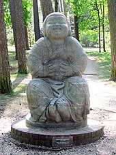 Frontale Farbfotografie eines Buddhas in Steinskulptur, der auf einem runden Sockel mit dem Schild „Bernhard Hoetger. Bonze des Humors 1914“ steht. Der Buddha hält sich lachend seinen dicken Bauch und hat seine Füße aneinander gepresst. Im Hintergrund sind Bäume und Waldwege.