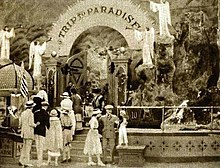 In the foreground, a man, a woman and a child are standing. Beside them a queue of people entering through a portal whose header reads "trip to paradise".