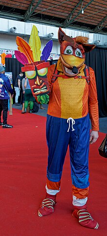 Photo d'un homme dans un costume de marsupial orange et un pantalon bleu, avec un masque vaudou porté à la main droite.