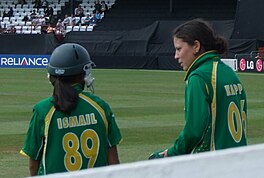 Two female in green cricket uniforms with yellow piping are talking. The left-hand female is wearing a helmet and has dark skin, 'ISMAIL' and '89' are visible in yellow writing on her back. The right-hand female has 'KAPP' and '06' visible on her back.