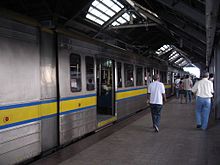 A metallic metro train with a blue yellow and blue stripe running along its side waits with doors open at a station with a dark roof and flaps letting in some sunlight as disembarked passengers head for the exit