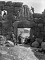 Schliemann schaut am berühmten Löwentor von Mykene durch ein Loch in der Mauer.