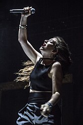 Lorde wears a black crop top and skirt as she raises her arm and tilts her head upward with her eyes closed. A sole spotlight shines on her.