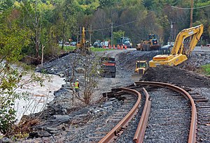 Construction equipment and workers in a stony depression at the side of a body of water on the left. Bent railroad tracks dangle into it from the foreground; traffic is backed up on a road in the rear