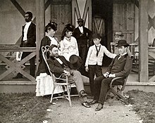 Photographie de famille prise sur le perron d'une maison en bois. Grant en costume et avec un haut-de-forme à la main est assis dans une chaise en osier face à un jeune homme assis dans une chaise pliable en bois et portant un chapeau melon. Derrière eux se trouvent deux femmes en robe et deux garçons.