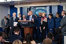 Trump vorbește în sala de briefing din aripa de vest, cu diverși oficiali în picioare în spatele lui, toți îmbrăcați în ținute oficiale și fără măști de față