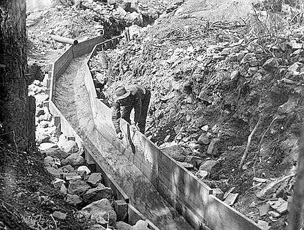 A man leans over a wooden sluice. Rocks line the outside of the wood boards that create the sluice.