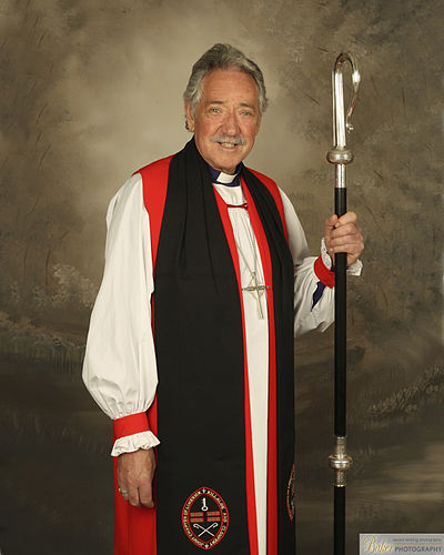 An Anglican bishop in choir dress: purple cassock, rochet, red chimere and cuffs, tippet, and pectoral cross.