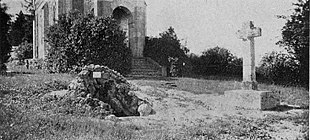Photographie en noir et blanc représentant une croix et un tertre placés devant un bâtiment religieux.
