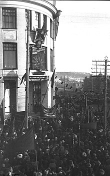 Photographie den noir et blanc d'une rue pleine de manifestants agitants des drapeaux communistes devant un bâtiment pavoisé d'un drapeau communiste et avec l'étoile rouge où se trouve la faucille et le marteau dessus.