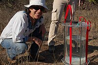 Woman wearing a white hat, white shirt, and blue jeans squatting in dry lands next to a water container.
