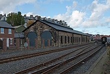 A long stone built building with an inverted V-shaped roof, with railway lines in the foreground
