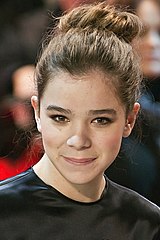 Ein Bild eines lächelnden hellhäutigen Teenager-Mädchens mit ihrer Hand auf ihrer Hüfte. Sie hat langes hellbraunes Haar und trägt ein schwarzes Oberteil mit transparenten Ärmeln.