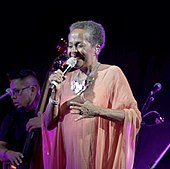 A woman in a blue dress holding a microphone with her right hand and smiling to the ground.