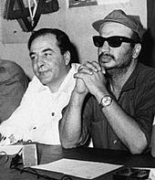 Dva sedící muži za stolem. Jeden je oblečen v bílé košili, druhý ve vojenské khaki košili s čepicí a černými brýlemi.