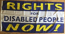 This flag has three horizontal stripes with colours and text. The stripes are blue, white and blue. The text on the blue stripes says "RIGHTS" and "NOW!" in yellow letters, and the text in the middle white stripe says "FOR DISABLED PEOPLE" in smaller print.