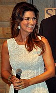 Shania Twain wearing a white dress is standing holding a microphone and smiling.